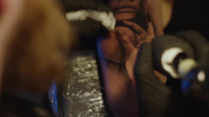 A tattoo artist carefully wipes away excess ink from a realistic black and grey portrait tattoo on a client's arm