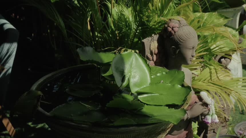 A traditional Thai statue stands in a serene garden next to a water bowl with lily pads