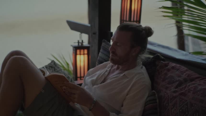 A tranquil man with a man bun relaxes on a patterned couch while reading a book in a dimly lit beach cabana at dusk