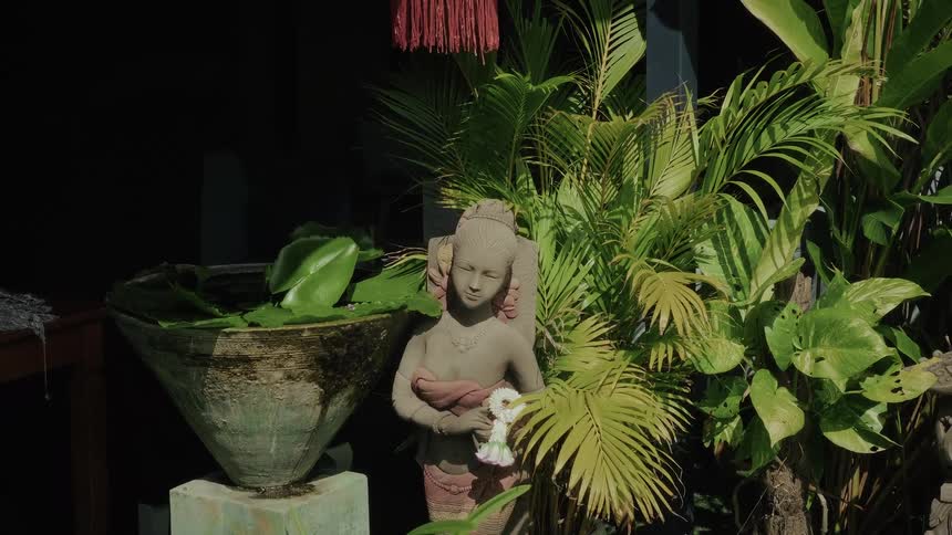 A stone statue of a woman holding a flower garland stands amidst lush tropical plants and a water basin