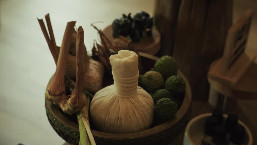 A close-up of a decorative bowl filled with a traditional Thai herbal compress, kaffir limes, and various spices for a spa treatment