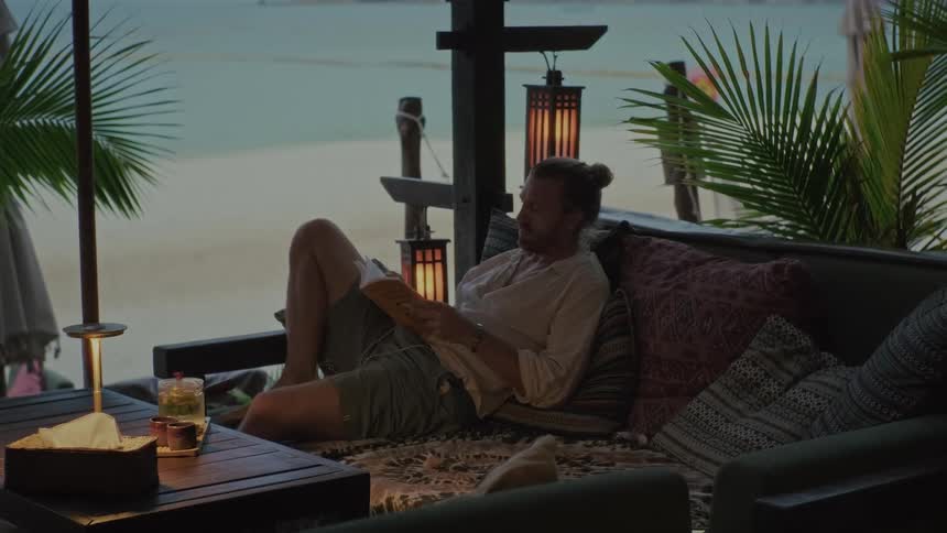 A man with his hair in a bun relaxes on a comfortable sofa reading a book in a cozy, dimly lit beachside lounge at dusk