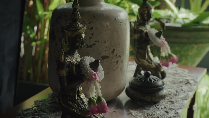 A close-up of traditional Thai deity statues adorned with floral garlands, resting on a table in a tranquil, sunlit room
