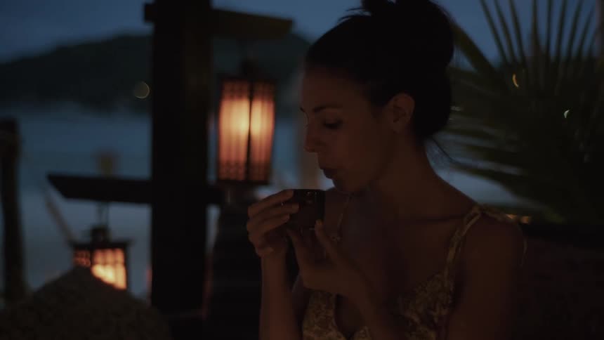A pensive woman sips tea at a tranquil beachside retreat at dusk, illuminated by the warm glow of lanterns