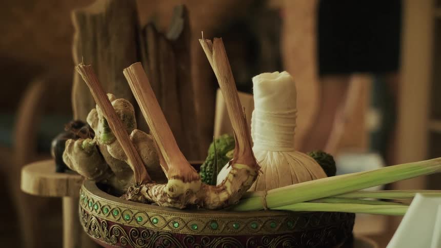 A close-up of a decorative bowl with natural Thai massage ingredients like galangal, ginger, and an herbal compress ball