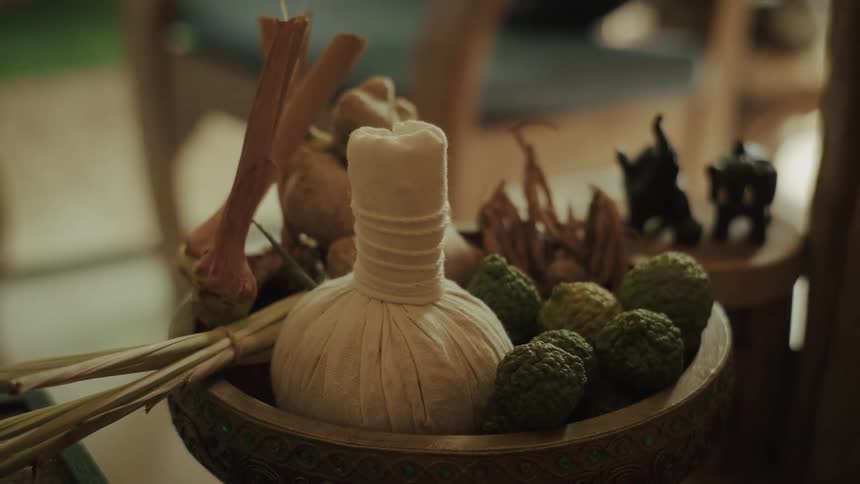 An ornate bowl holds a traditional Thai herbal compress ball, kaffir limes, lemongrass, and ginger for a relaxing spa treatment