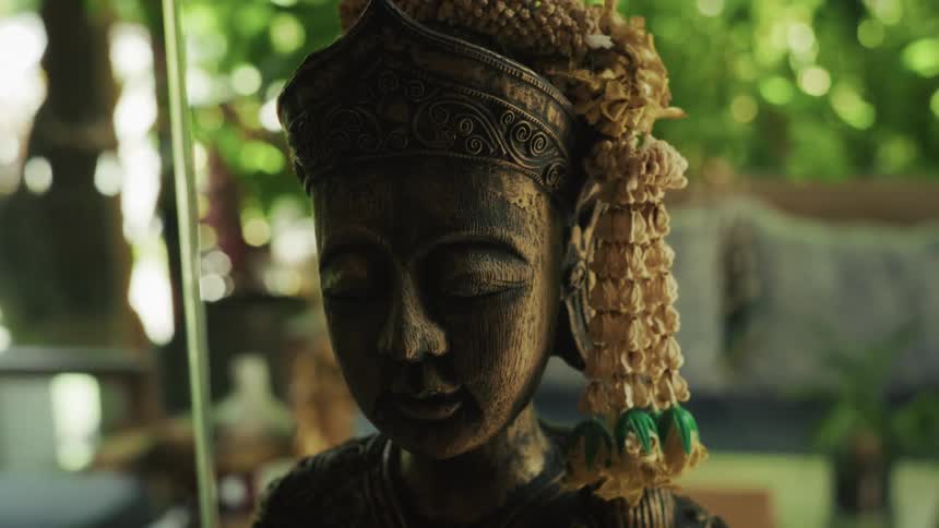 A meditative wooden statue with closed eyes and a dried flower garland sits peacefully in a lush green environment
