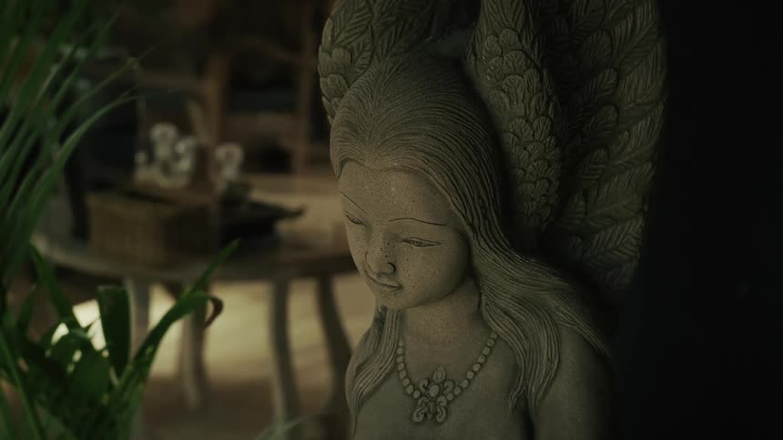 A serene stone sculpture of a female angel with detailed wings sits in a softly lit, tranquil room