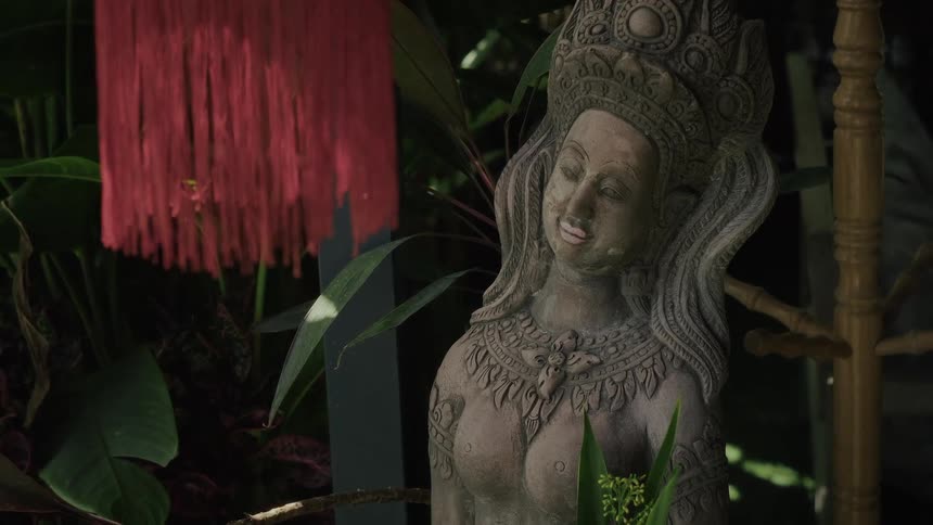 A serene stone statue of a goddess stands in a lush garden holding an offering of purple orchid flowers