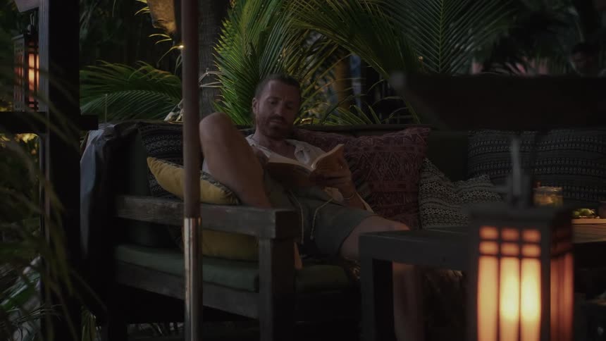 A man enjoys a peaceful evening reading a book by lantern light on a comfortable outdoor sofa at a tropical resort