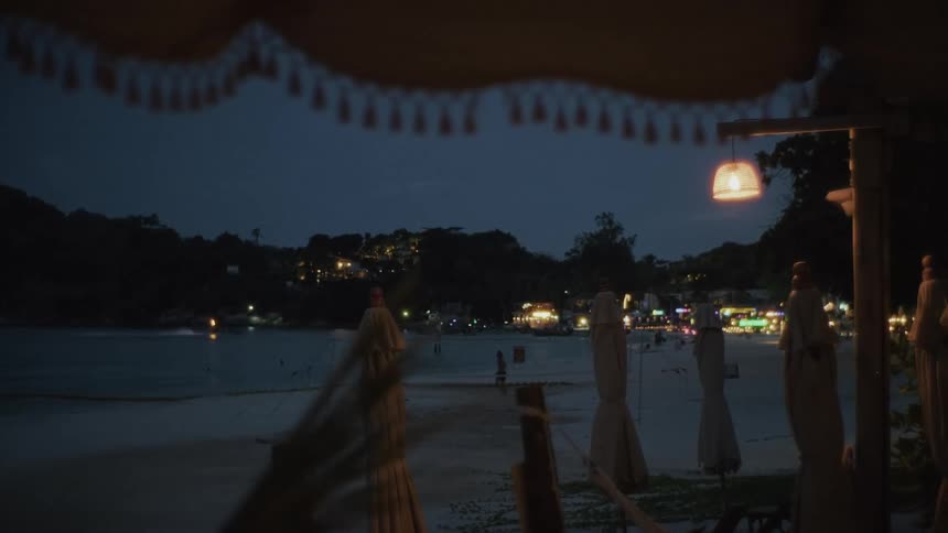 A tranquil evening view from a beach bar shows a tropical beach at dusk with distant lights from a hillside town