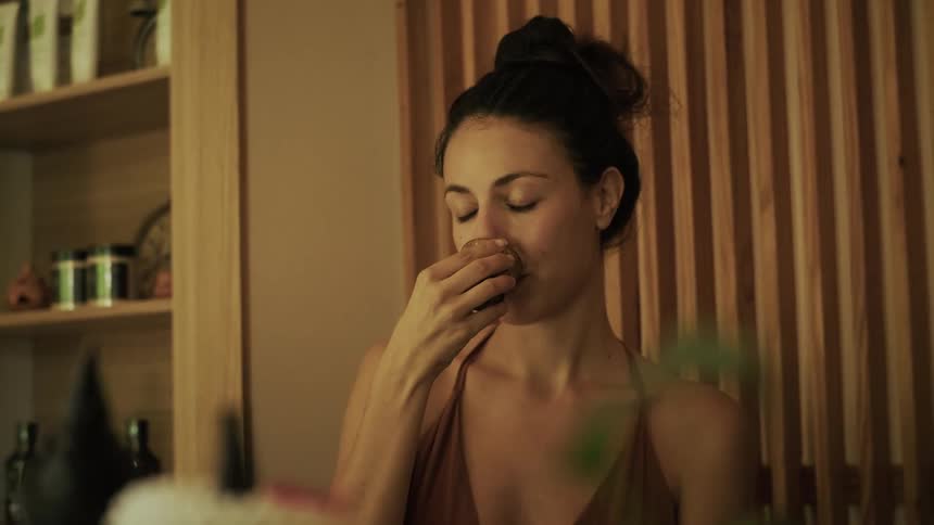 A serene woman with her eyes closed mindfully smells and drinks tea from a traditional cup in slow motion