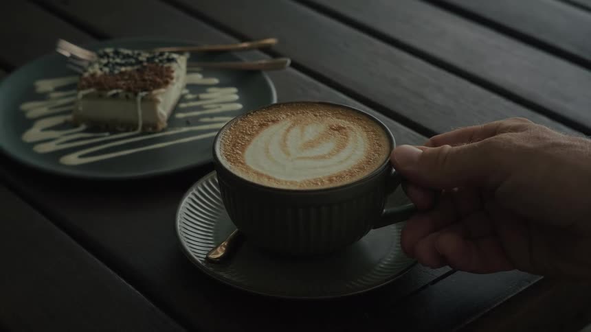 A man's hand slowly picks up a cup of freshly made cappuccino with latte art from a dark wooden table in a cafe