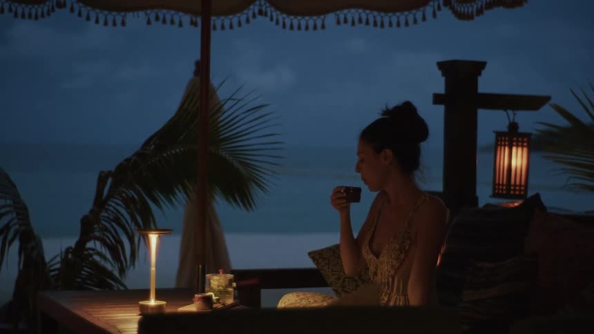 A woman enjoys a peaceful evening alone, sipping from a cup at a softly lit table in a tropical beachfront restaurant at dusk