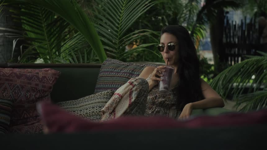 A woman with sunglasses relaxes on a couch in a tropical lounge and sips a smoothie in slow motion