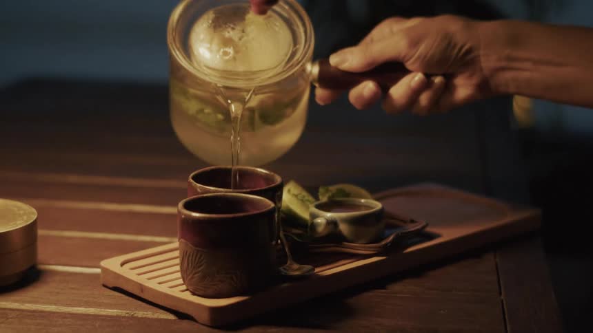 A slow-motion close-up captures a hand gently pouring hot tea from a glass pot into a traditional cup for a serene tea ceremony at night