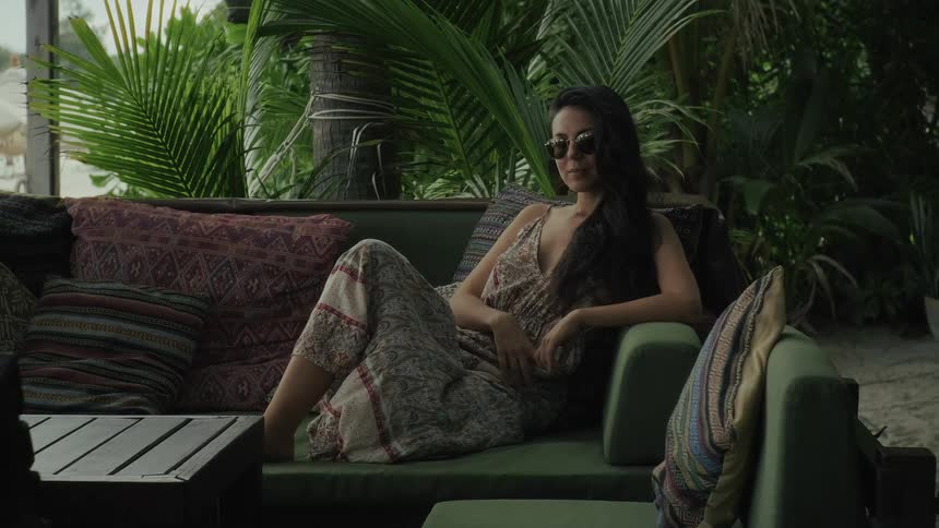 A stylish woman in sunglasses relaxes on an outdoor sofa at a tropical resort surrounded by lush palm trees