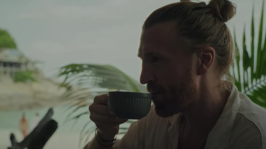 Slow-motion close-up of a contemplative man with a beard and hair bun mindfully sipping a drink at a tropical beach resort