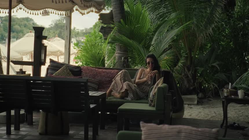 A woman relaxes on a plush couch at a tropical beachside resort, sipping a drink and enjoying the serene atmosphere