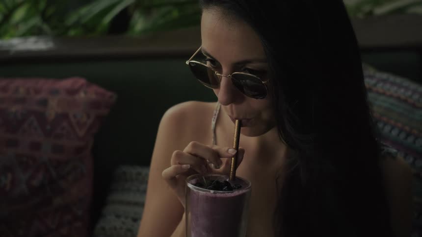 A beautiful woman in sunglasses slowly drinks a purple fruit smoothie through a straw and smiles