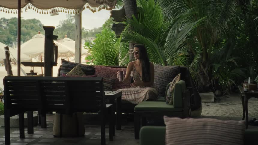A stylish woman in sunglasses relaxes with a drink on a plush couch at a tropical beachside lounge