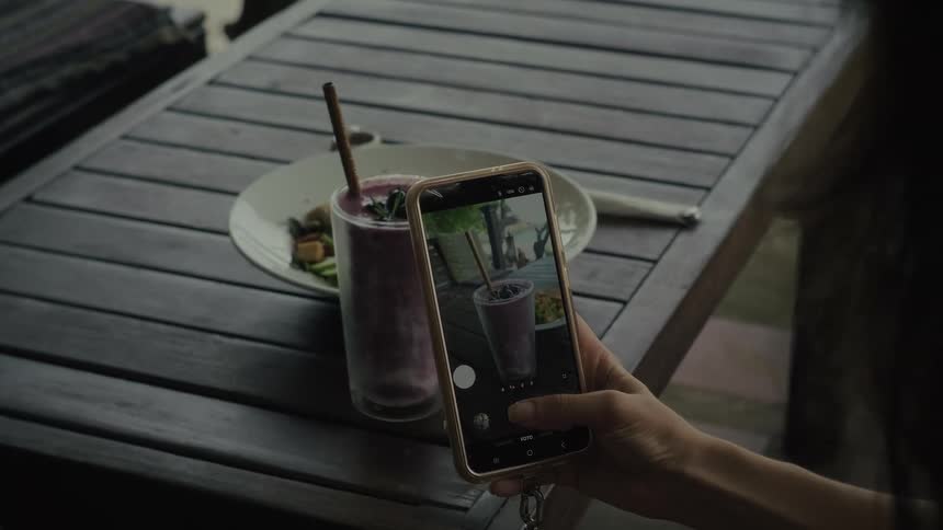 A person uses their smartphone to take a picture of a healthy purple smoothie and a salad on an outdoor wooden table