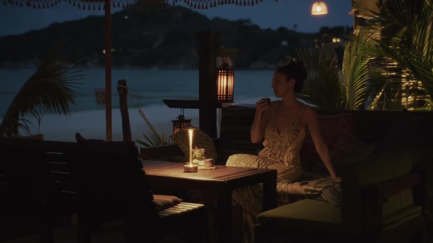 A woman relaxes at a dimly lit beachside restaurant, pouring and sipping tea by candlelight with the ocean in the background