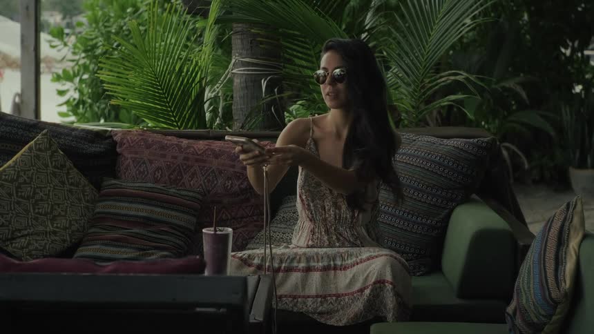 A stylish young woman in a sundress takes selfies on her phone while relaxing on a comfortable couch in a tropical lounge