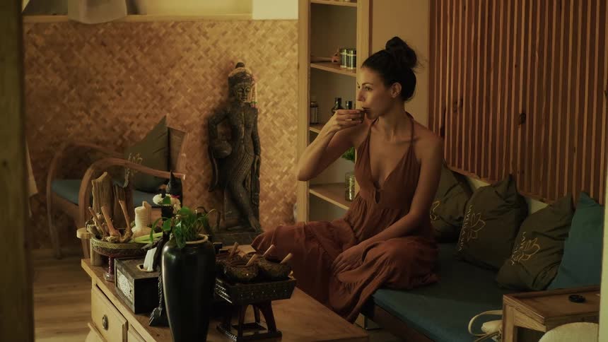 A woman in a long dress relaxes in a serene, Asian-inspired room, slowly preparing and sipping a cup of herbal tea