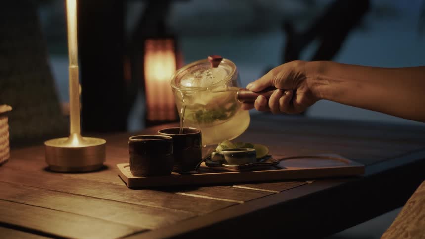 In a warmly lit, tranquil evening setting, a hand slowly pours hot herbal tea from a glass teapot into a ceramic cup