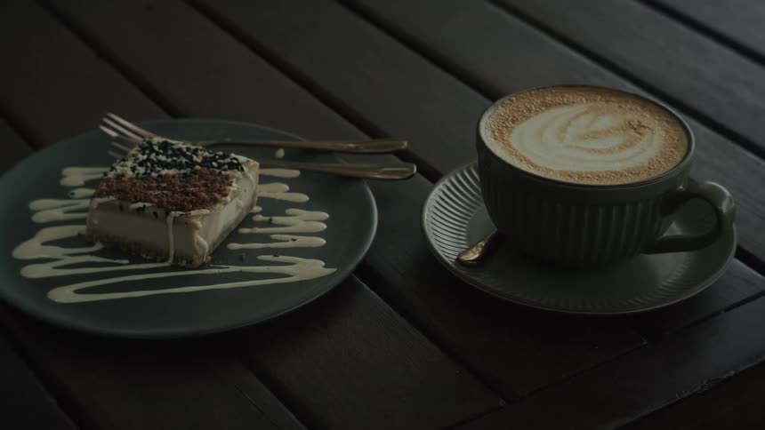 A hand picks up a cup of cappuccino with latte art sitting next to a slice of cheesecake on a dark wooden table in a cafe