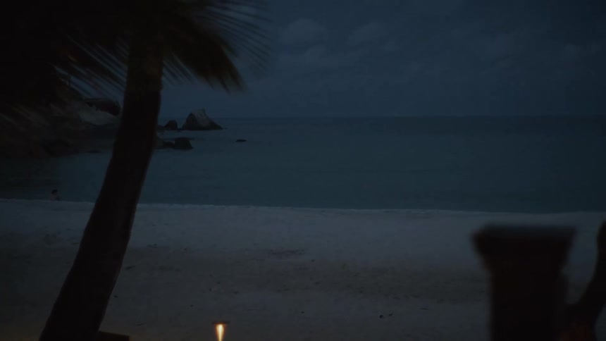 Quiet, candlelit tables for two sit on a sandy beach overlooking the gentle ocean waves at night