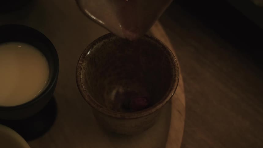 Slow-motion close-up of rose tea being poured into a rustic ceramic cup over a dried rosebud