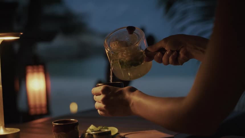 A person pours hot mint tea into a ceramic cup at a softly lit table during a peaceful evening outdoors