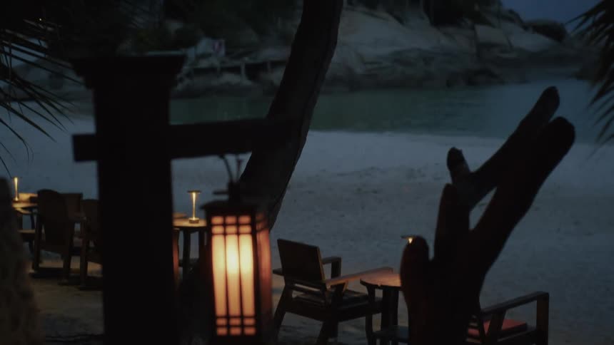 An empty beachside restaurant is illuminated by glowing lanterns during a calm evening as the camera reveals the sandy shore and a quiet bay