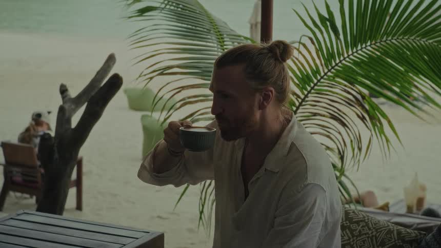 A man with his hair in a bun slowly sips from a cup of coffee while relaxing at a tranquil beach cafe