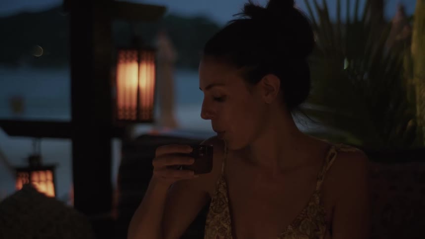 A woman enjoys a peaceful moment while sipping a beverage from a small cup at a lantern-lit beachside restaurant at dusk
