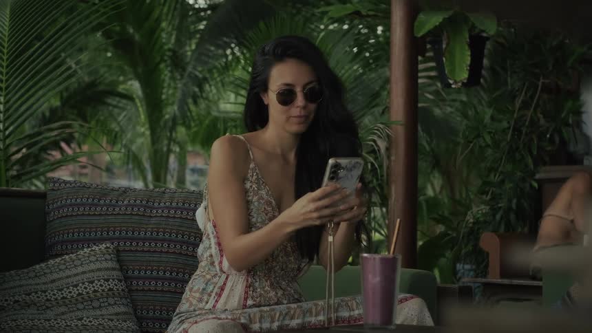 A chic woman in sunglasses relaxes at a tropical cafe, taking a selfie while enjoying a smoothie