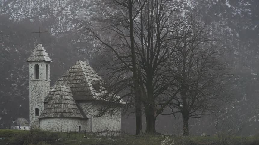 A historic stone church with a wooden shingle roof stands on a desolate hill in a foggy mountain landscape