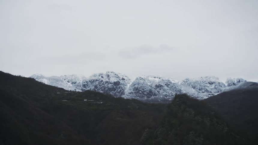 A slow camera pan across a dark mountain range reveals a distant, majestic snow-capped mountain range under a cloudy sky