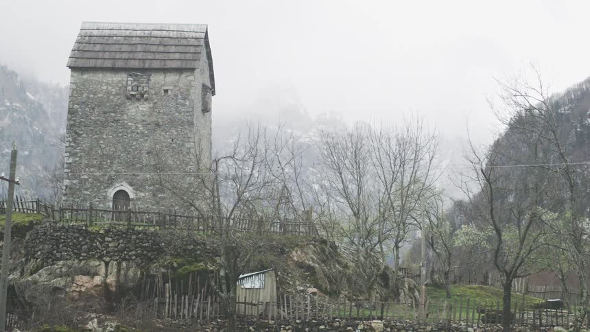 A historic stone watchtower stands on a small hill in a remote mountain village on a foggy, overcast day