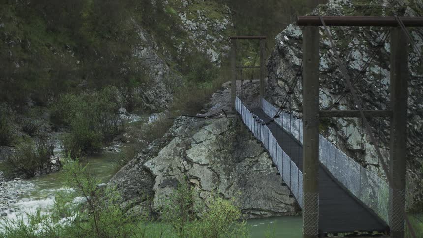 A static shot shows a remote suspension bridge spanning a flowing river in a rugged, green mountain canyon