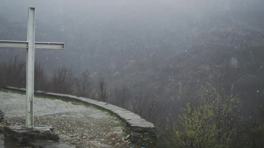 A lone Christian cross stands on a mountainside viewpoint as heavy snow falls over the foggy, desolate valley below
