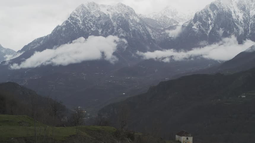 A vast, moody valley sits below towering snow-capped mountains partially obscured by clouds on a gray day