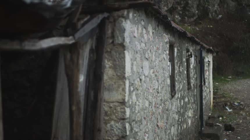Rain falls in slow motion on a weathered and abandoned stone building with a rusty tin roof in a desolate, rocky landscape