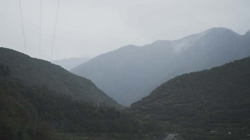 An atmospheric view of a winding road and rocky river through a lush mountain valley on a rainy and foggy day
