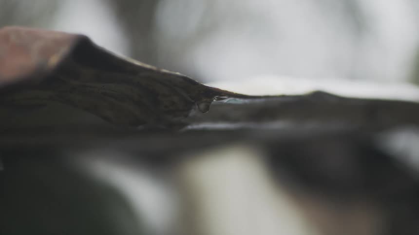 A close-up slow-motion shot of raindrops forming and dripping from the edge of a rustic tin roof