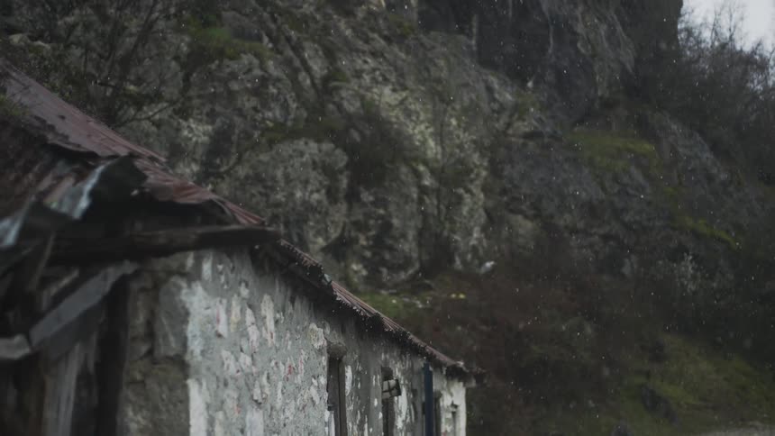 Snowflakes gently fall on a rustic, abandoned stone building with a rusty roof at the base of a mossy cliff