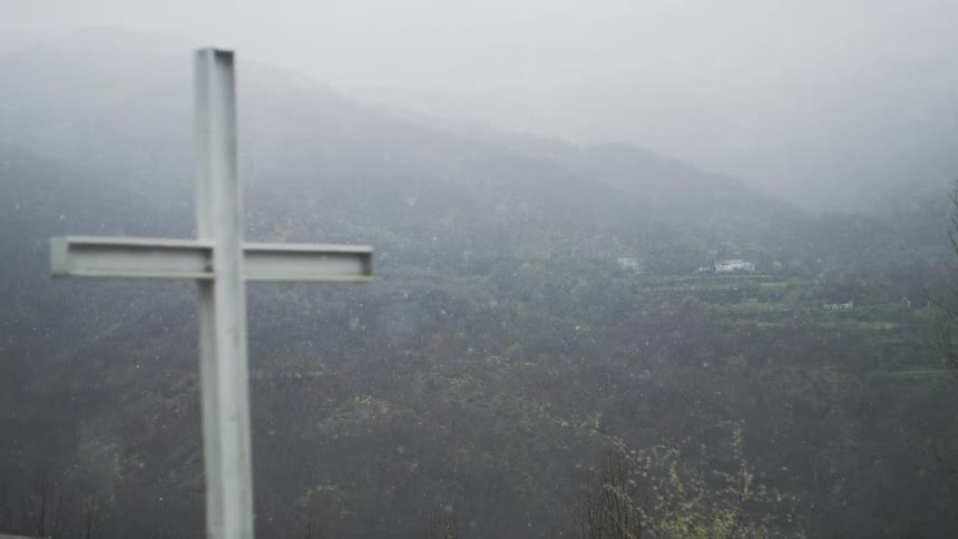 A Christian cross stands on a hilltop overlooking a valley as snow gently falls in slow motion