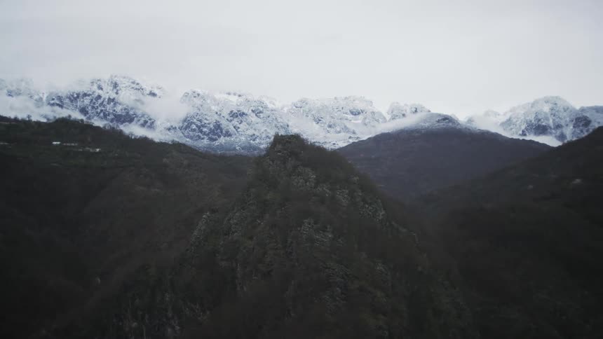 A slow aerial pan across a vast and moody landscape, revealing dark, rugged hills with snow-capped mountains in the distance